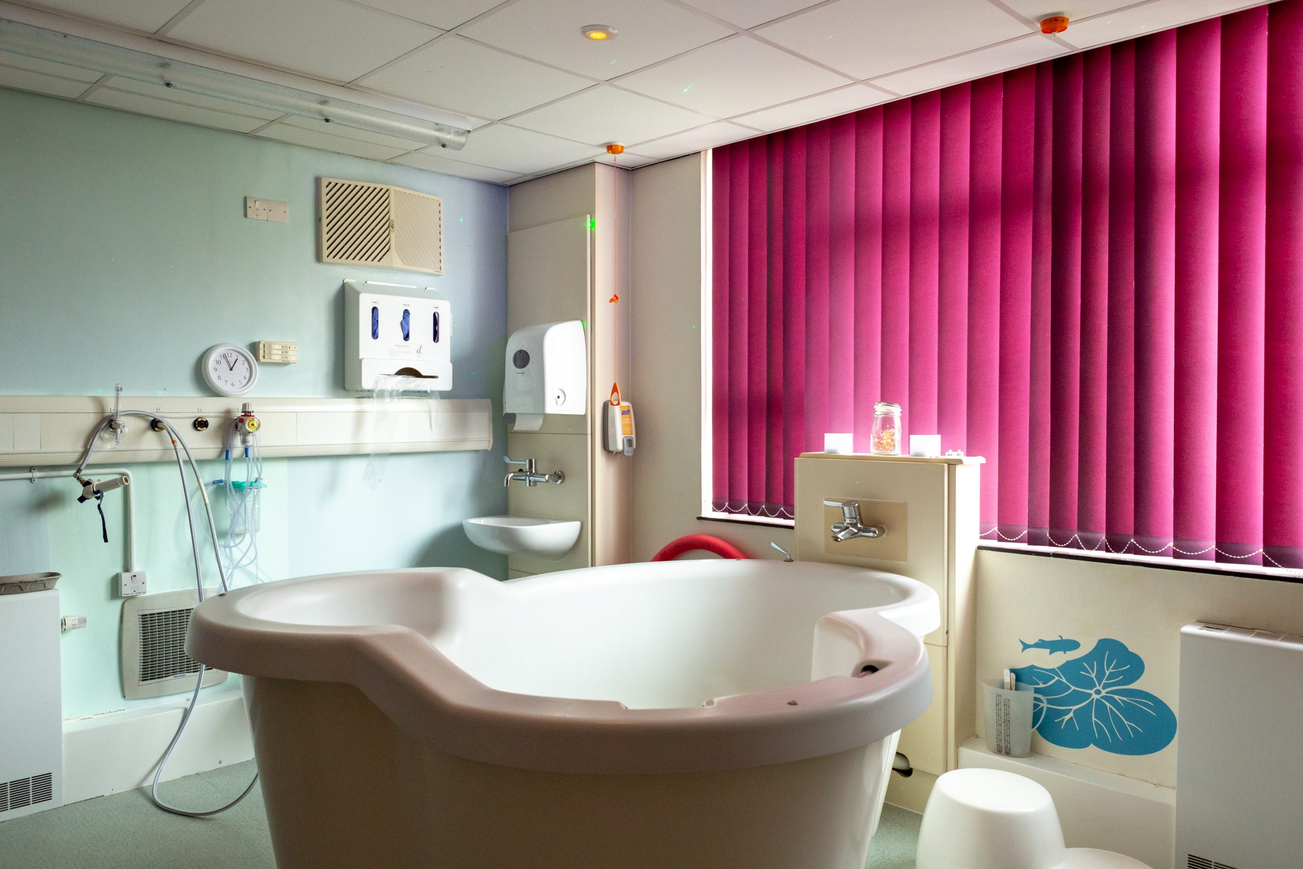 This image shows a hospital birthing room with a large bathtub in the middle of the room and a water tap near the edge of the bathtub. The room walls are painted a light blue colour and on the left side of the wall are a disposable glove dispenser, a clock, a sink and paper towel dispenser. The right side of the wall has large pink blinds that are closed but still allow some light into the room.