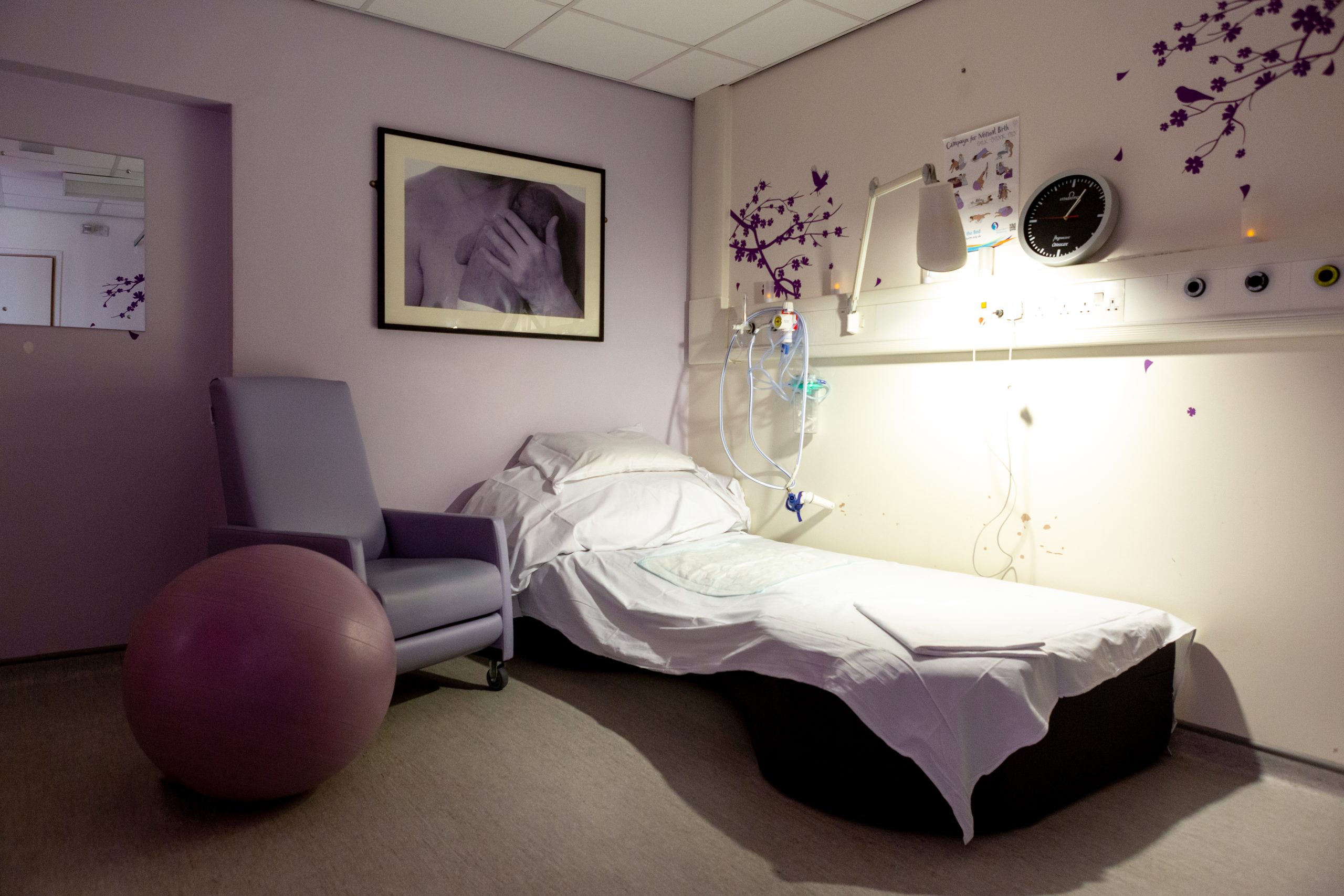 The image shows a softly lit hospital room with a hospital bed neatly made with white sheets and positioned against the wall. On the left of the bed is an armchair and a large exercise resting nearby the armchair. The walls are painted a light purple lavender colour with a dark purple branch tree and flower design. Above the bed is large black photo frame with a black and white image of a mother holding their newborn baby’s hand.