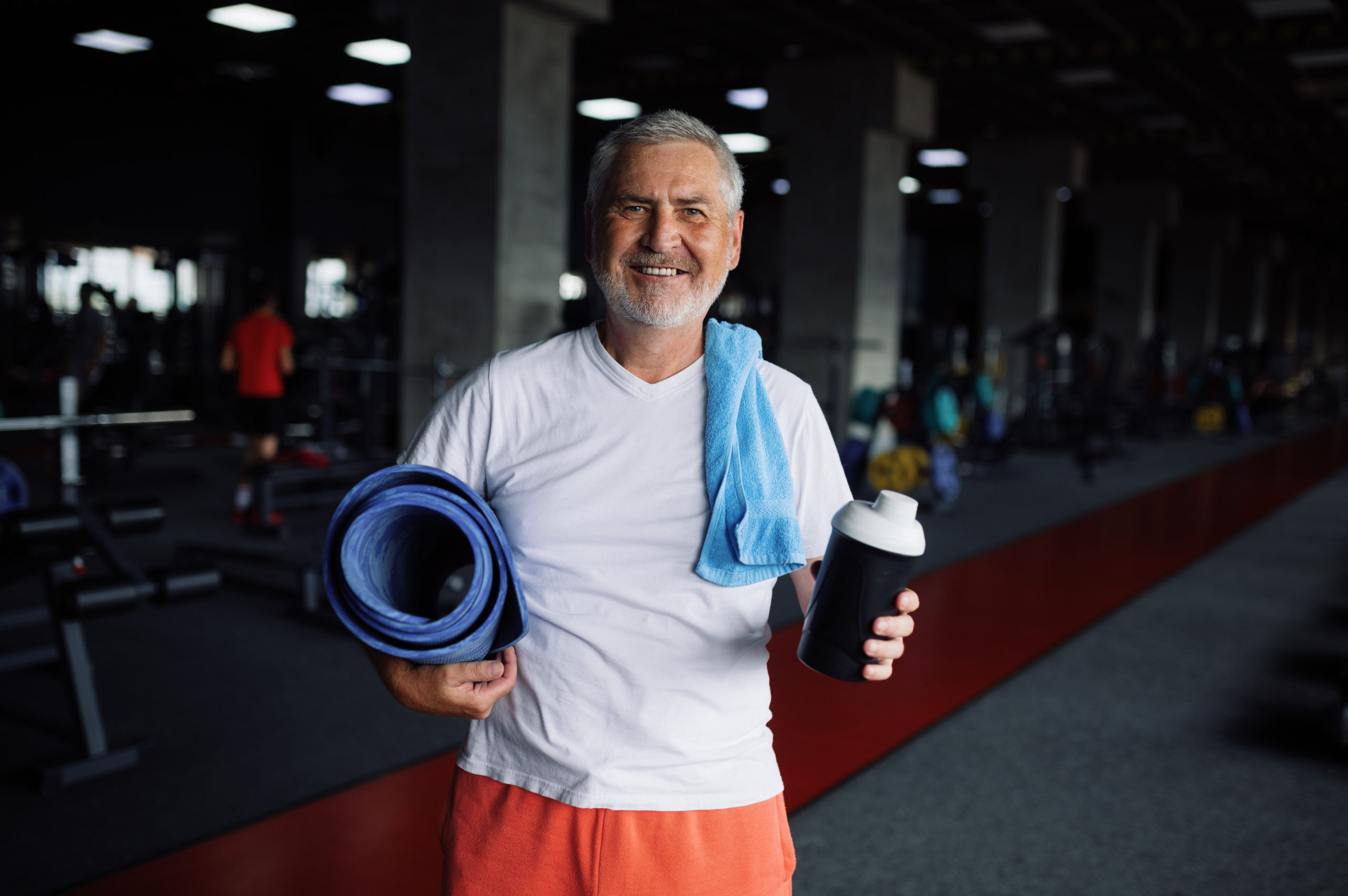 Happy old man with bottle of water, towel and mat, gym interior on background. Sportive grandpa on fitness training in sport center. Healty lifestyle, health care, elderly sportsman