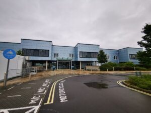 An image showing the front of Bridgwater hospital. The hospital is a blue fronted building with the first floor sections protruding over the ground floor level. These level one extensions also have glass spanning the whole part of the section.