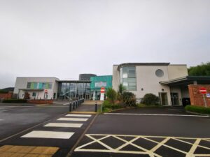 A photo of the front of Minehead Community Hospital. The right of the building is a off white colour with a curved glass going towards a cooper green fronted entrance. This is connected to a glass building with a stairs. To the left of that is a off white building. All these sections of the building are attached.