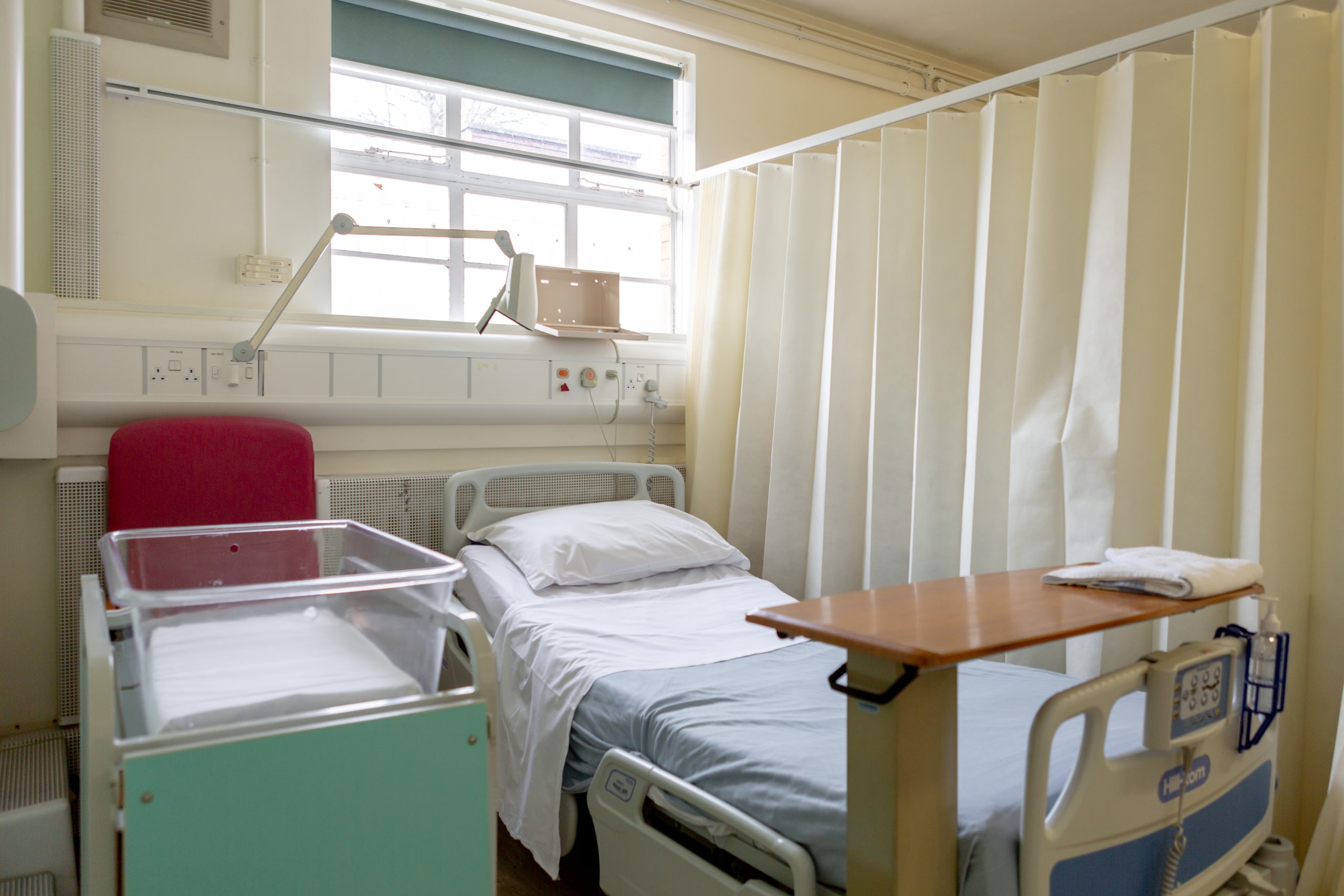 A well organised and clean hospital space with a single bed. The bed has a single white pillow with light blue sheets, a moveable wooden table is attached to the bottom of the bed. A red armchair is positioned next to the bed. Above the bed are various medical and electrical outlets. The wall is painted a light cream colour with a green roller shade above the head of the bed.