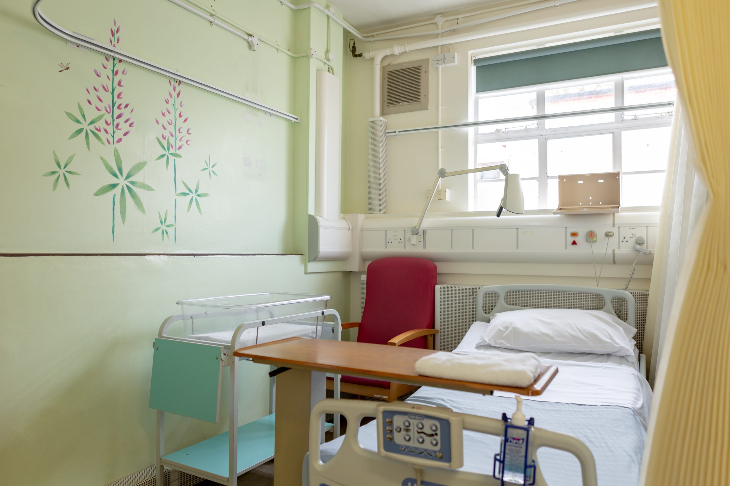 A hospital room with a single bed. The bed has a white pillow and light blue sheets with a moveable wooden table towards the bottom of the bed. A red armchair is located next to the head of the bed. The wall is painted light green and has a decorative mural of pink and green flowers. The room has various medical equipment associated with the maternity ward and to prepare for a new mother and her baby.