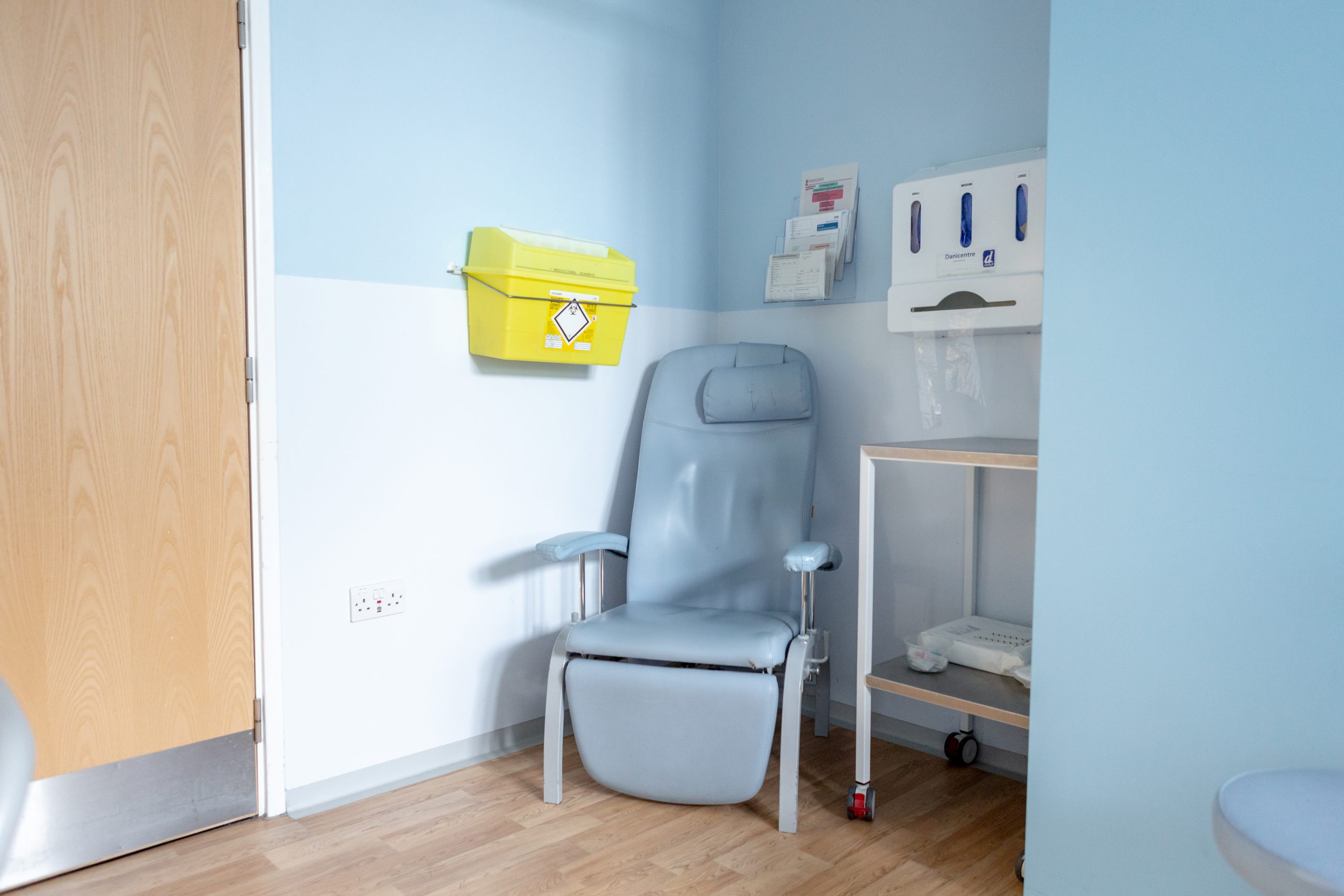 The image shows a light blue armchair placed in the corner of a room with a yellow medical waste bin mounted on the left wall. Mounted on the right wall is a dispenser containing disposable gloves and leaflet shelves containing various printed handouts. The room is painted light blue with a two-tone design and with wooden flooring.