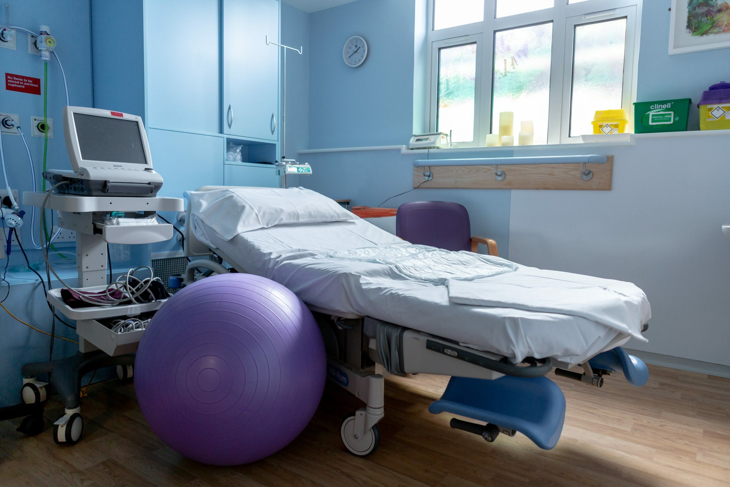 The image shows a hospital room with a neatly made bed with white sheets. To the left of the bed is a large purple exercise ball and a monitor on a wheeled cart with various cables and wires attached to the monitor. The room has light blue walls, cabinets and a window letting natural light in.