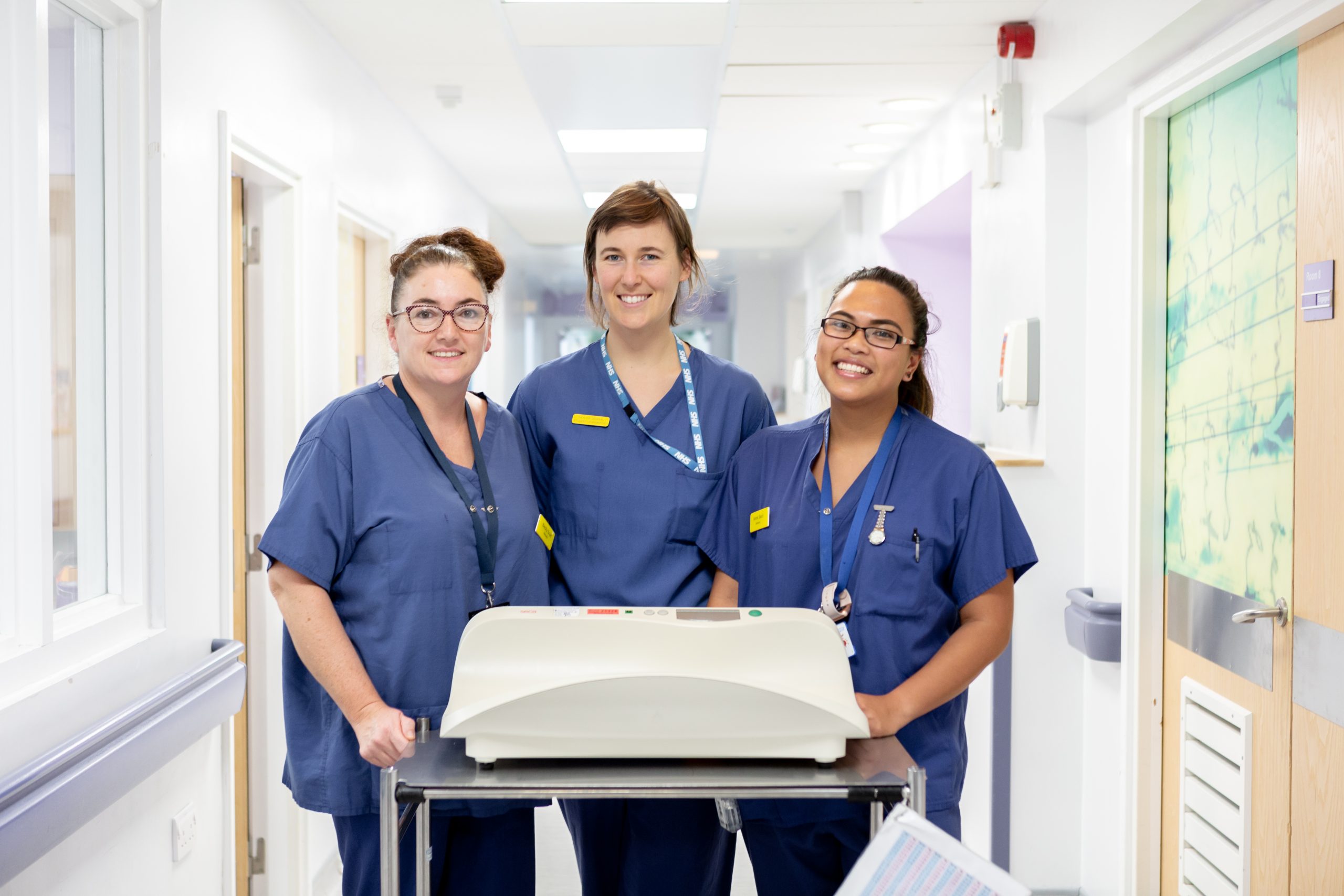 The image shows three Trust colleagues dressed in dark navy-blue scrubs standing behind a medical equipment in a ward hallway with bright smiles on their faces.