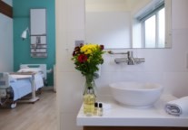 The image features a hotel-style white worktop with white porcelain sink resting on top. The front right corner of the worktop has a small rolled towel and to the left is a small vase of fresh flowers and small bottles of toiletries. Above the sink, a brushed silver tap is attached to the wall and simple sleek rectangular mirror can be seen higher up, reflecting the bathroom window. Through the doorway you can see a neatly made up hospital bed with lamp over.
