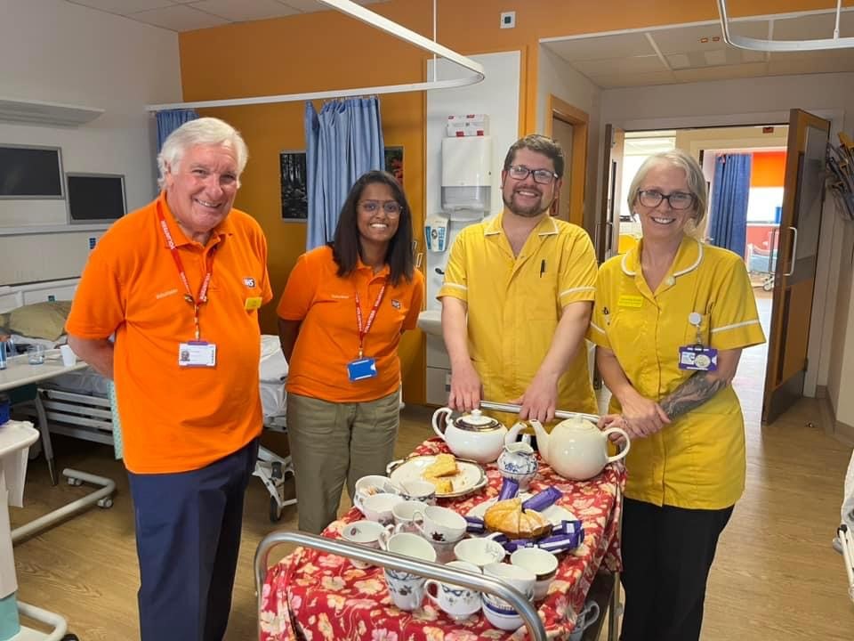 Volunteers stood round a trolley with tea cups, tea pots, cake and mini rolls on a plate. They are stood in a ward setting with curtains behind them.