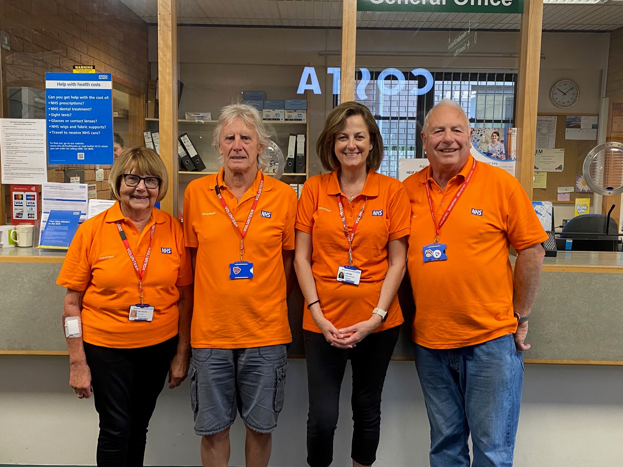 Four volunteers stood in front of the general office at Musgrove Park Hospital. they are all wearing orange with red volunteer lanyards.