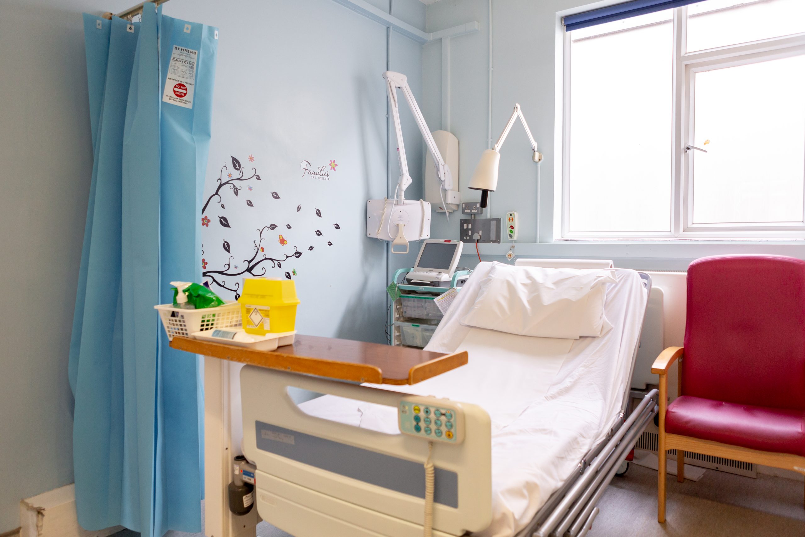 A hospital space with a single raised bed with a white pillow and a moveable wooden table at the bottom of the bed with various medical equipment around the bed. A red armchair is next to the bed. The wall is painted a light blue colour with a small mural of trees and leaves.